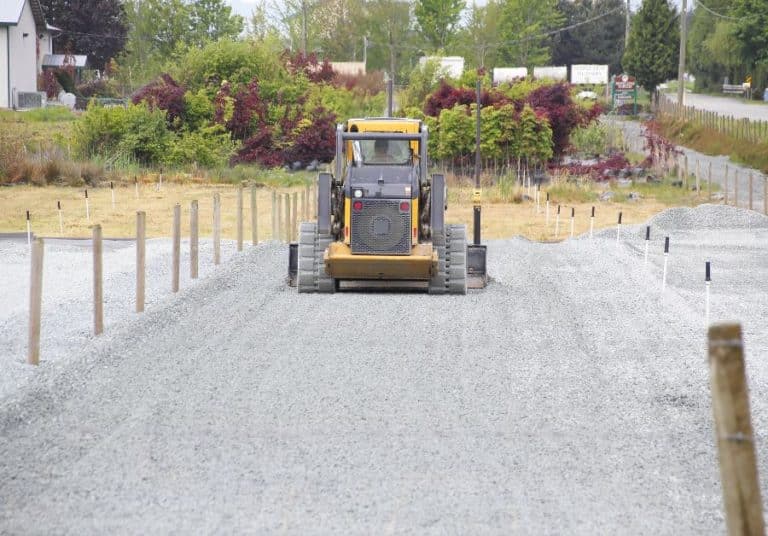 A yellow bulldozer leveling a roadbed of gray gravel with wooden stakes lining the path and greenery in the background.
