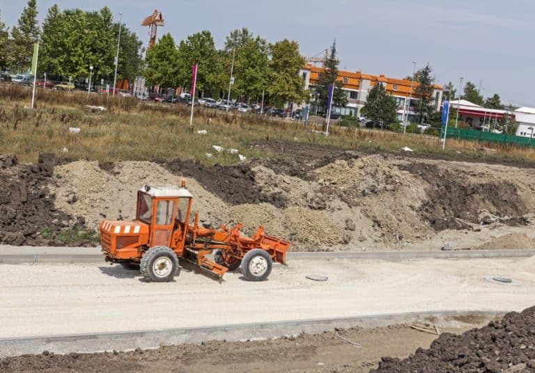 Orange grader on a construction site with a freshly leveled roadbed, dirt piles, and urban background.