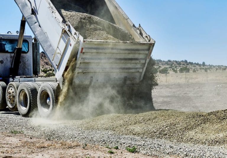 A heavy-duty dump truck unloads a large pile of gravel, creating a cloud of dust against a sparse landscape under a clear sky.