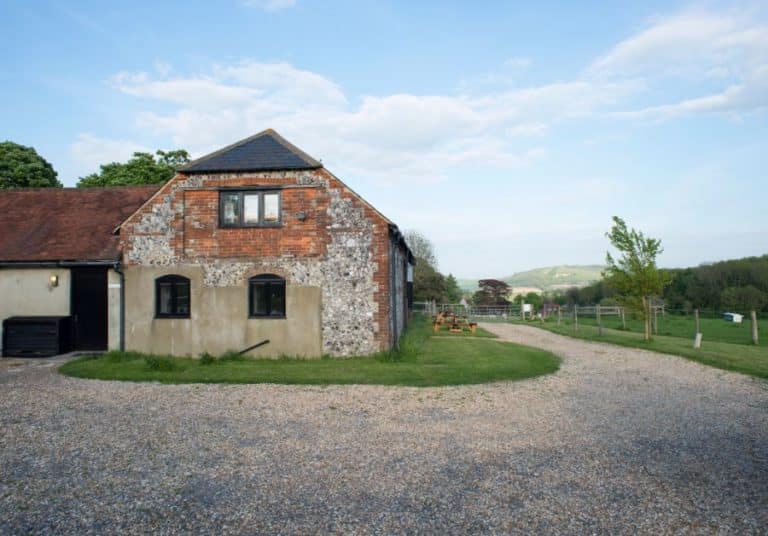 Renovated old brick farmhouse with exposed flint walls, featuring a gravel driveway, set against a serene rural landscape with rolling hills in the distance.