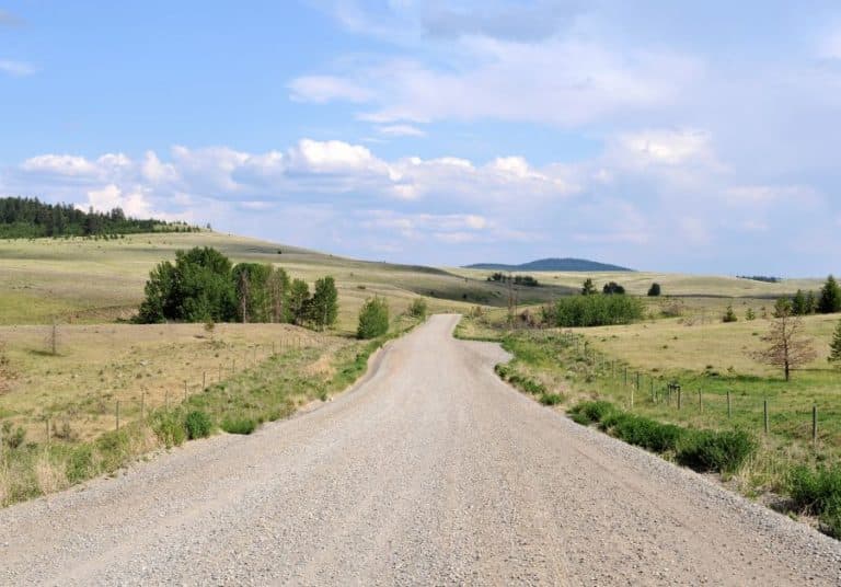 Open gravel road meandering through rolling hills and open fields under a blue sky with scattered clouds.