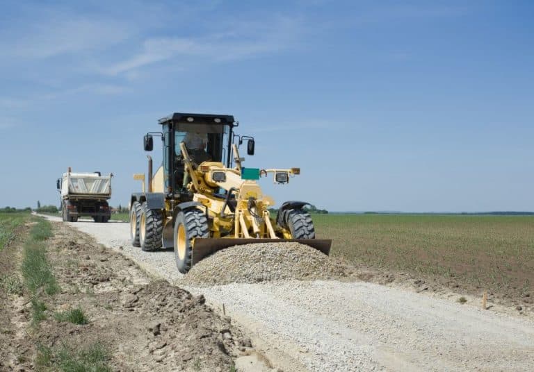 Yellow motor grader leveling gravel on road construction site under clear sky.