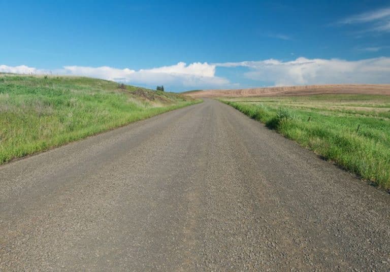 Rural gravel road meandering through a lush green landscape under a clear blue sky with distant rolling hills.